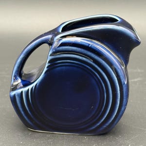 May include: A small, deep cobalt blue ceramic creamer pitcher featuring a rounded body with concentric ring detailing. The pitcher has a curved handle on the left side and a prominent pouring spout on the right, with a glossy, reflective finish.