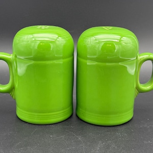 May include: A pair of lime green ceramic salt and pepper shakers, each featuring a cylindrical body, a rounded top, and a single side handle. The shakers have a smooth, glossy finish with subtle horizontal ring detailing near the base.