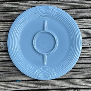 May include: A light blue, round ceramic serving platter with a central dip area and three divided sections. The platter has a concentric ring design and decorative arch details. The plate is on a weathered wooden surface.