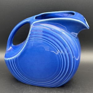 May include: A cobalt blue ceramic pitcher featuring a rounded, disc-like body with concentric circular embossed rings on the side. The vessel includes a curved handle on the left side and a prominent, pointed pouring spout on the right side.
