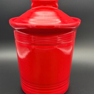 May include: A bright red ceramic canister with a matching lid. The cylindrical container features horizontal ringed detailing near the top and bottom edges. The lid has a smooth, domed shape with a small, rectangular handle at the very top.