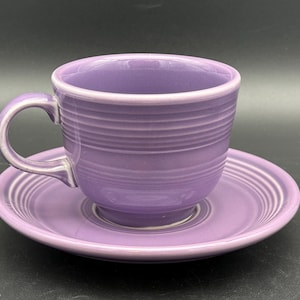 May include: A lavender ceramic tea cup and matching saucer set. The cup features a rounded handle and horizontal ribbed detailing around the exterior. The saucer has a matching circular ribbed pattern along the outer edge, creating a cohesive look.