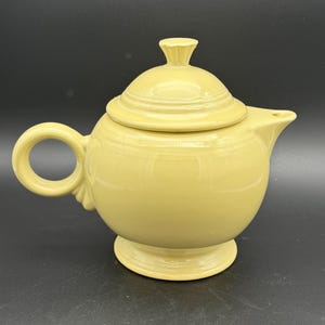 May include: A pale yellow ceramic teapot with a lid and a rounded body. The teapot has a handle, a spout, and sits on a small base. The lid has a small knob on top. The teapot is a solid color and has a glossy finish.