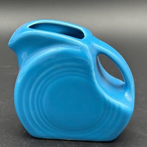 May include: A turquoise ceramic pitcher with a rounded body and a curved handle. The pitcher has a spout for pouring and concentric circles on the front. The bright blue color and unique shape make it a decorative item.