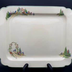 May include: A rectangular cream-colored ceramic serving platter with scalloped edges and a thin green border. The platter features colorful painted garden scenes in the corners, including a stone path, a floral archway, and trees.