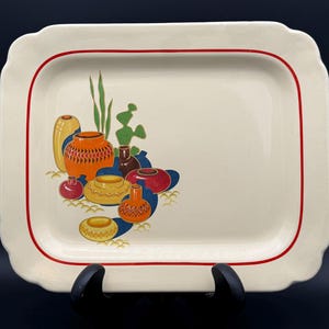 May include: A rectangular cream-colored ceramic serving platter with scalloped edges and red trim. The center features a colorful graphic illustration of various small pottery vessels in shades of orange, yellow, red, and blue, accented by green foliage.