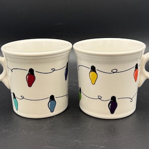 May include: Two off-white ceramic mugs featuring a repeating pattern of black string lights with colorful bulbs in red, blue, yellow, green, and purple. Each mug has a round handle and a smooth, glossy finish, displaying a festive holiday theme.