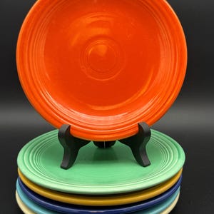 May include: A stack of five round ceramic plates featuring concentric ring patterns along the rims. The top plate is bright orange, followed by light green, yellow, dark blue, and light blue plates. A black plastic stand holds the orange plate.