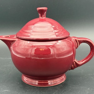 May include: A deep red ceramic teapot with a glossy finish, featuring a rounded body, a matching lid with a small knob handle, a curved spout, and a side handle. The design includes subtle ribbed detailing at the base of the spout and handle.