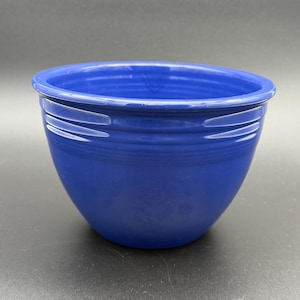 May include: A deep cobalt blue ceramic bowl featuring a smooth, glossy finish and a series of three horizontal concentric rings embossed around the upper exterior rim. The bowl has a wide, rounded opening and a tapered base, showcasing a solid color.