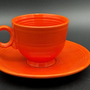 May include: A vibrant orange teacup and saucer set. The cup has a rounded shape with a circular handle. The saucer is round with a slightly raised edge. Both pieces have a glossy finish and are made of ceramic.