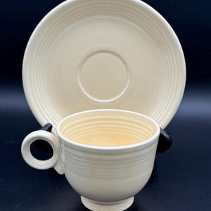 May include: Cream-colored teacup and saucer set. The cup has a rounded shape with a handle and horizontal ridged detailing. The saucer is round with concentric ridges. The set is made of ceramic.