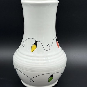 May include: White ceramic vase with a festive design. The vase features a series of colorful Christmas lights in yellow, red, and green, connected by a black line. The vase has a bulbous base and a slightly flared top.