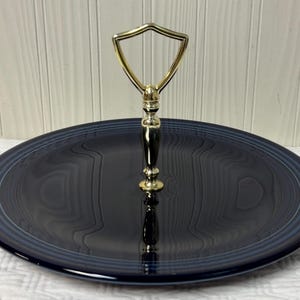 May include: A navy blue serving platter with a gold-toned handle. The round platter has a glossy finish and a raised rim with a blue accent. The handle is shield-shaped.
