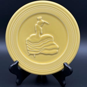 May include: A yellow ceramic plate with a raised relief design of a dancer in a ruffled dress. The plate has a ridged border and is displayed on a black stand. The plate is a light yellow color.