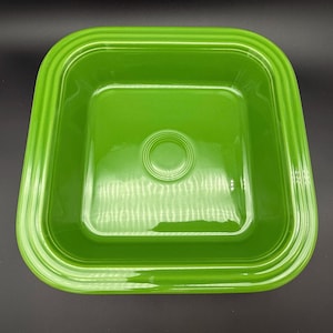 May include: A vibrant green square ceramic serving dish. The dish has a glossy finish and a slightly raised rim. The center features a circular indentation. The overall design is simple and modern, suitable for serving food.