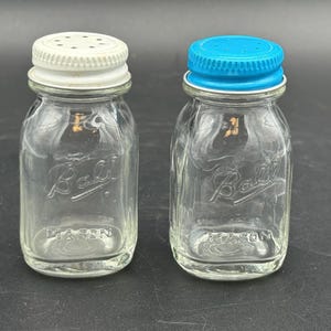 Puede incluir: Dos saleros de vidrio transparente con el logotipo Ball Mason en relieve en el frente. Uno tiene una tapa de rosca de metal blanco con peque&ntilde;os orificios, mientras que el otro tiene una tapa de rosca de metal azul brillante con orificios.