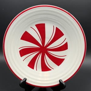 May include: A round white ceramic plate featuring a central red peppermint swirl pattern and a thin red rim. The plate has a slightly raised edge and a smooth, glossy finish, displaying a classic circular design reminiscent of a holiday candy.