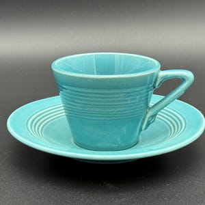 May include: A turquoise ceramic teacup and saucer set. The cup has a handle and horizontal ridged detailing around the body. The saucer also has ridged detailing. The set is a solid, bright turquoise color.