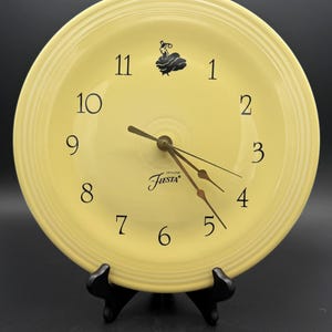 May include: A round, pale yellow ceramic wall clock featuring concentric ring detailing on the rim. The clock face displays black Arabic numerals from 1 to 12, a black silhouette graphic of a person, and the text Genuine Fiesta in black script.