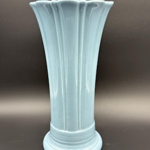 May include: A light blue ceramic vase with a fluted design. The vase has a wide, scalloped rim and a round base. The vertical fluting gives the vase a textured appearance. The vase is approximately 8 inches tall.