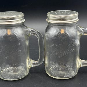 May include: Two clear glass mason jar mugs with handles and silver-colored metal shaker lids. Each jar features the embossed Ball logo and the word MASON on the front. The lids have small perforated holes for dispensing contents.