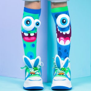 May include: A pair of novelty knee-high socks with monster faces and matching sneakers. The socks are green and blue with large cartoon eyes and open mouths. The sneakers are white, blue, and green with bunny ear accents and a "Toy Story" charm.