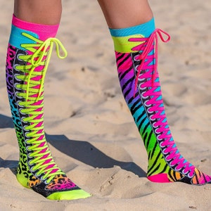Puede incluir: Un par de calcetines altos de colores con un diseño de cordones. Los calcetines presentan un patrón de color rosa brillante, amarillo, azul y verde con un estampado de animales en blanco y negro. Los calcetines se llevan en las piernas de una persona y están de pie sobre una superficie arenosa.