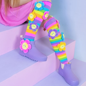 May include: Purple rain boots with a rainbow striped sock featuring colorful flower appliques with smiley faces.