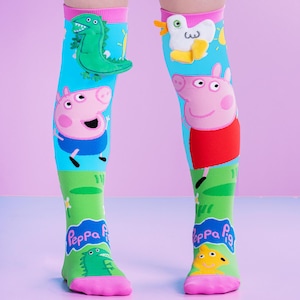 Pode incluir: Meias de cano alto com personagens da Peppa Pig. As meias são principalmente turquesas, com detalhes verdes, rosa e amarelos. Uma meia tem um dinossauro verde em 3D, a outra um pato em 3D. As meias também apresentam a Peppa Pig e outros personagens.