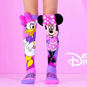 May include: Knee-high socks featuring Daisy Duck and Minnie Mouse. The socks are pink and purple with cartoon graphics of the characters. The Disney logo is visible in the background.
