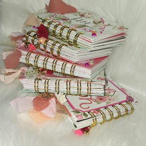 May include: A stack of floral-themed spiral notebooks with gold-colored binding. The covers feature pink and white floral patterns, with decorative ribbons and charms. Some notebooks have text on the cover. The notebooks are stacked on a white, fluffy surface.
