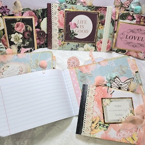 May include: A collection of handmade journals and notebooks with floral and butterfly designs. The journals feature decorative lace, ribbons, and embellishments. Some have text that reads "Life is Good", "Lovely", and "make memories today".