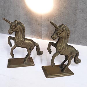 May include: A pair of matching brass unicorn figurines, each depicted in a rearing pose with a single spiralled horn on its forehead. The unicorns are mounted on rectangular brass bases and feature a metallic, aged gold-toned finish.