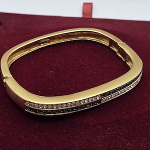 May include: A gold-toned hinged bangle bracelet with a rectangular shape. The front section features two parallel rows of embedded stones, with one row of clear white crystals and one row of dark blue square-cut stones set into the metal band.