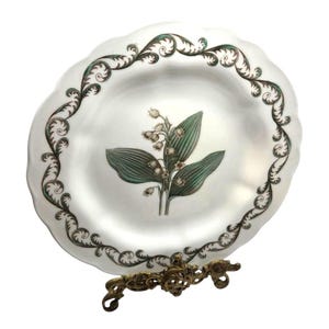 May include: A white ceramic plate with a scalloped edge featuring a central illustration of a lily of the valley plant with green leaves and white flowers, surrounded by a dark green and black swirling foliate border pattern on the rim.