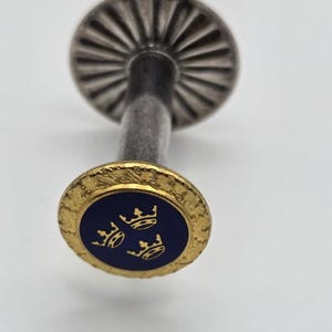 May include: A metal stick pin featuring a circular head with a dark blue enamel centre displaying three gold-coloured crowns. The head is surrounded by a textured gold-tone border, and the pin includes a silver-toned fluted disc component.