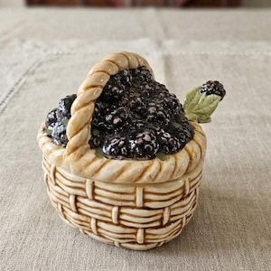 May include: A ceramic lidded jar shaped like a woven basket filled with dark purple blackberries. The basket has a light tan finish with a braided handle. A small green leaf and a single blackberry are attached to the side of the lid as a handle.