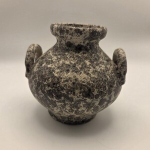 May include: A small, round ceramic vase featuring a textured, speckled finish in shades of dark grey and off-white. The vessel has a wide, short neck with a flared rim and two small, curved handles attached to opposite sides of its body.
