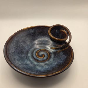 May include: A round ceramic dish with a dark blue and brown glaze. The interior features a spiral pattern embossed in the centre, and a curved, handle-like spiral element extends from the rim towards the middle of the bowl.