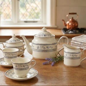 May include: A cream-coloured porcelain tea set featuring a delicate blue floral pattern and silver trim. The set includes a teapot with a lid, a sugar bowl with a lid, a creamer, and two teacups with matching saucers, all arranged on a wooden surface.