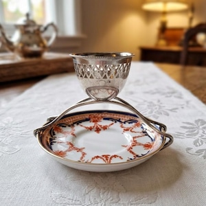 Peut inclure: Un coquetier en métal argenté avec un bord ajouré décoratif, soutenu par deux bras métalliques incurvés fixés sur une petite soucoupe en porcelaine blanche ornée d'un motif floral détaillé dans les tons orange et bleu marine.