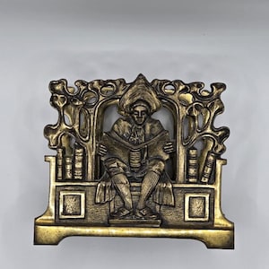 May include: A brass-coloured metal napkin holder featuring a relief design of a person wearing a hat and sitting on a bench while reading a book. The background includes stylised tree branches and stacks of books on either side of the central figure.