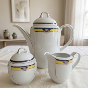 May include: A white porcelain coffee service set consisting of a tall coffee pot with a lid, a lidded sugar bowl, and a creamer. Each piece features a horizontal band of yellow, dark blue, and lavender accents with thin gold trim detailing.