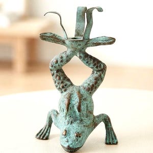 May include: A metal candle holder shaped like a frog standing on its front legs in a handstand position. The frog has a textured, weathered teal and brown patina finish. A circular candle cup is positioned between its hind legs at the top.
