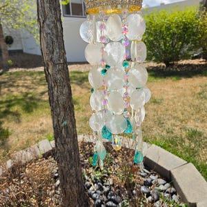 May include: A decorative wind chime featuring a gold-colored crown with clear and blue accents. The chime has iridescent, round, white shell-like discs and colorful beads. Teal and iridescent crystal-like teardrops hang from the bottom.