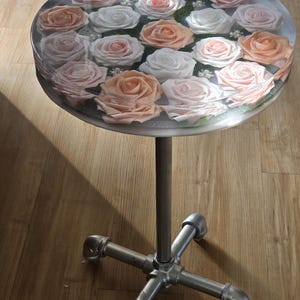 May include: A round table with a clear resin top filled with pink and white roses. The table has a silver metal base with a cross-shaped design.