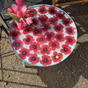 May include: A round, light blue table with red flower decorations and a vase of pink flowers. The table has a light blue base and is outdoors on a gravel surface. A few small stickers are on the table.