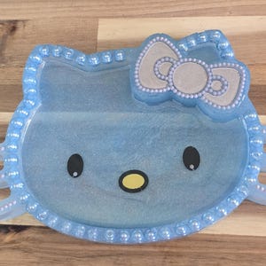 May include: A light blue resin tray in the shape of a cartoon cat face. The tray features a bow, eyes, and nose. The edges are lined with small, pearl-like beads. The tray is approximately 8 inches wide.