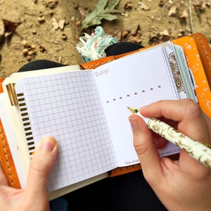 Könnte beinhalten: Ein offenes orangefarbenes Lederjournal mit einer karierten Seite und einer Seite mit dem Wort "Today." und einer Reihe von Punkten. Ein weißer Stift mit goldenem Blumendesign schreibt auf die Seite. Das Journal wird von einer Person offen gehalten.